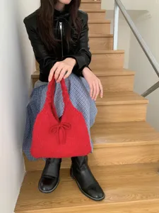 Lifestyle shot of red knitted handbag with bow detail held by model on wooden stairs.