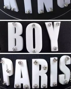 Dettaglio macro di lettere BOY PARIS in acrilico bianco con rivetti su cappello hip-hop nero.