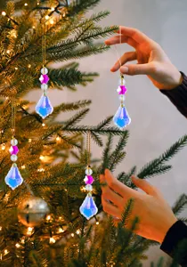 Hand hängt einen schillernden Kristall-Tropfenanhänger an einen beleuchteten Tannenbaum.