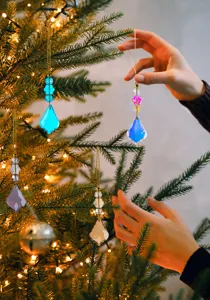 Handen die kleurrijke gefacetteerde kristallen hangers aan een verlichte kerstboom hangen.