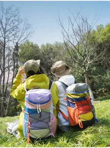 Δύο πεζοπόροι με πολύχρωμα σακίδια ξεκουράζονται σε έναν καταπράσινο λόφο στην εξοχή.