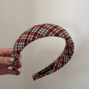 Détail d'un serre-tête à carreaux rouge et gris tenu à la main avec étiquette.