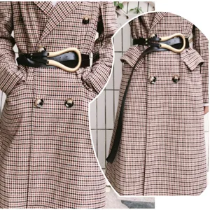 Détail de ceinture noire femme avec grande boucle dorée en U sur un manteau à carreaux.