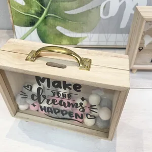 Detail view of a wooden piggy bank with 'Make YOUR dreams HAPPEN' and felt balls
