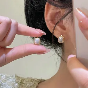 Close-up shot showing elegant square pearl earrings worn on an ear and held by a hand.