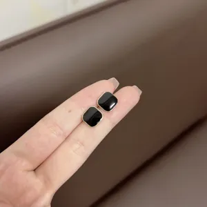 Detail view of square black enamel stud earrings with gold trim held on fingers.