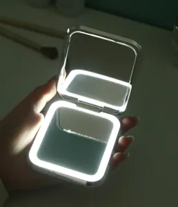 Détail d'une main tenant un miroir de maquillage pliable à LED argenté et portable