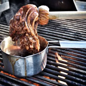 Macro detail view of a cotton BBQ basting mop dipping into a sauce pot over an outdoor grill.