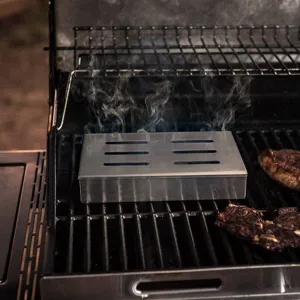 Stainless steel smoker box emitting smoke on a grill with meat for outdoor BBQ.