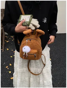 Mochila de peluche marrón con orejas de conejo y un pompón blanco sobre el modelo.