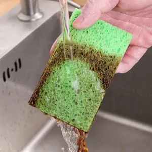 Green wood pulp cotton cleaning sponge being rinsed under water in a kitchen sink