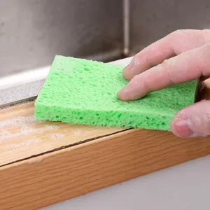 Green cellulose wood pulp sponge scrubbing a wooden surface in a kitchen sink setting.
