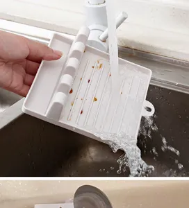 Close-up detail of white plastic kitchen spoon rest and pot lid holder being rinsed under sink faucet.