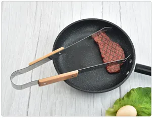 Detail view of stainless steel BBQ tongs with wooden handles flipping steak in a frying pan.