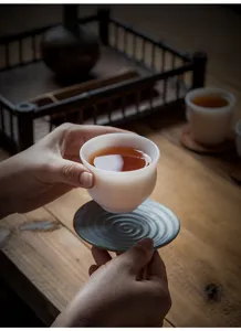 Hands holding a white Yangzhi jade porcelain Kung Fu tea cup with tea on a textured saucer.