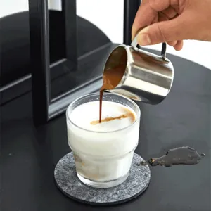 Nahaufnahme eines Edelstahl-Milchkännchens, das Espresso in cremigen Milchschaum gießt.