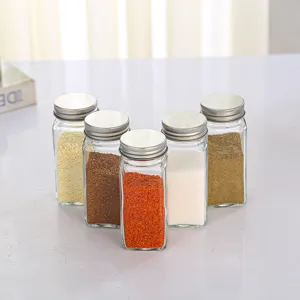 Vue détaillée de pots à épices carrés en verre avec couvercles argentés sur une table blanche.