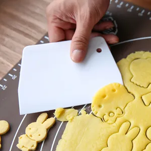 Gros plan d'un grattoir à pâte blanc en plastique coupant de la pâte à biscuits sur un tapis.