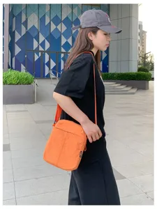 Modell trägt eine orangefarbene minimalistische Umhängetasche vor städtischem Hintergrund