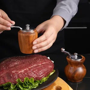 Acacia wood manual pepper grinder detail, hand cranking spices over beef on kitchen desk.