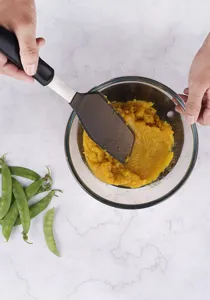 Detail shot of a black silicone spatula mixing food in a glass bowl on marble countertop.