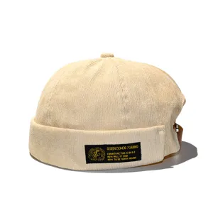 Detail shot of a beige corduroy brimless docker hat with a decorative black patch