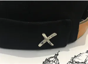 Detail shot of black brimless docker cap, featuring brown leather strap and metal cross emblem.