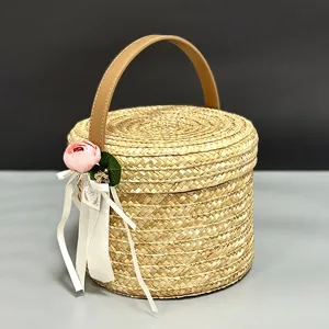 Cesta de regalo redonda de paja tejida a mano con asa de cuero y etiqueta de flor decorativa.