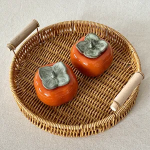 Vassoio rotondo in rattan intrecciato con manici in legno e due decori di cachi in ceramica.