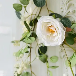 Close-up of white artificial rose on green eucalyptus vine against ornate white wall backdrop