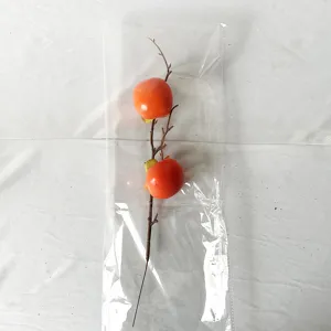Dettaglio di un ramo di cachi finto con due frutti arancioni in confezione di plastica.