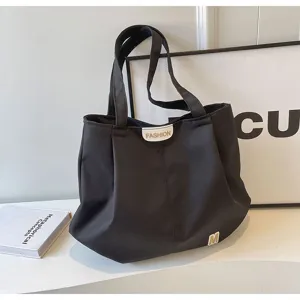 Detalle de bolso de hombro de lona negra con diseño plisado y etiqueta de letra frontal.