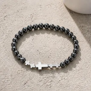 Primer plano de pulsera de hematita con dijes de cruz plateada sobre fondo de piedra.