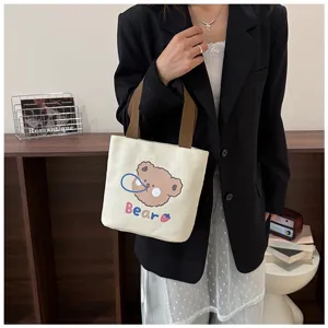 Detalle de un bolso de lona crema con un diseño de oso de dibujos animados y asas marrones.
