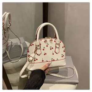 Detailansicht einer weißen Muschel-Handtasche mit süßen roten Kirschmotiven und Goldakzenten.