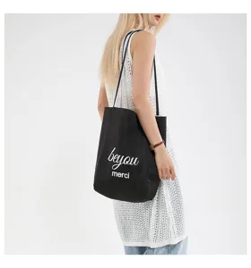 Modelo luciendo un bolso tote de lona negra con detalle de texto 'beyou merci' blanco