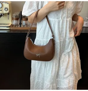 Bruine hobo schoudertas met zilveren strik, gedragen door een model in witte jurk
