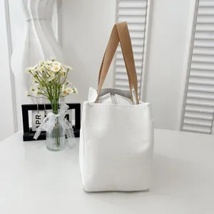 Side view of a minimalist white canvas lunch bag with tan handles on a white desk.