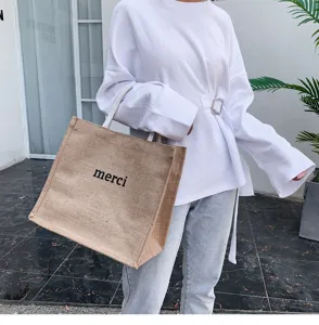 Detail shot of a large jute tote bag with merci print carried by a model in white.