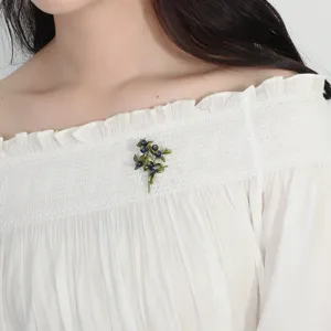 Détail d'une broche myrtille vintage avec feuilles vertes sur un chemisier blanc.