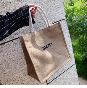 Close-up of burlap tote bag with black 'merci' print and white woven handles.