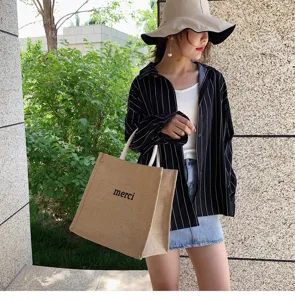 Casual large capacity jute tote bag with merci print held by model in an outdoor setting