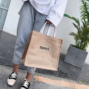 Detail view showing the merci print on a large capacity jute tote bag with white handles