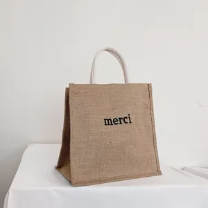 Detail shot of a rustic burlap tote bag featuring 'merci' print and white rope handles