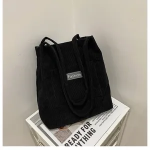 Schwarze Cord-Tragetasche mit Fashion-Patch auf weißem Regal-Display präsentiert.