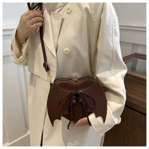 Detailweergave van bruine hartvormige vleermuisvleugel crossbodytas bij een beige trenchcoat.