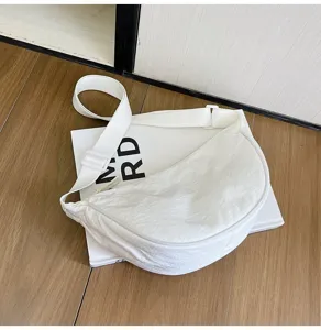 Bolso bandolera de nailon blanco minimalista en forma de media luna sobre libro y suelo.