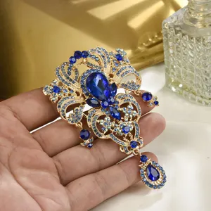 Detailansicht einer verzierten Goldbrosche mit leuchtend blauen Tropfenkristallen in der Hand.