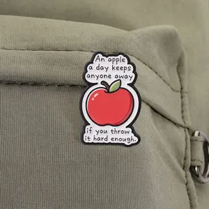 Close-up of a funny red apple enamel pin with humorous English text on a green backpack