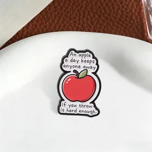 Close-up of funny red apple enamel pin with humorous English quote on white plate.